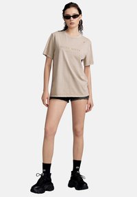 Camiseta beige de gran tamaño con detalles desgastados y letras que dicen "MISS SEXY". Llevada con pantalones cortos negros y zapatillas negras gruesas. Los accesorios incluyen gafas de sol.