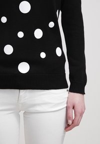 Czarny dziergany sweter w białe kropki z prążkowanymi mankietami i dolnym brzegiem. Częściowo widoczne białe jeansy. Teksturowana tkanina.