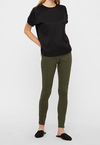 Top nero con dettagli in pizzo sulle spalle, abbinato a pantaloni verde oliva attillati e scarpe slip-on nere. Tessuto liscio con silhouette casual.