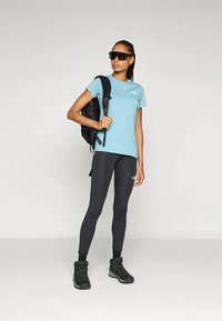 Šviesiai mėlyna trumpomis rankovėmis marškinėliai, juodos tamprės, juodi žygio batai ir juoda kuprinė, tamsūs akiniai nuo saulės ir prigludęs dizainas.