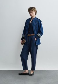 Kvinde iført en blå denimjakke, marineblå bukser, sorte ballerinasko og holder en brun taske, står op ad en ensfarvet lys væg.