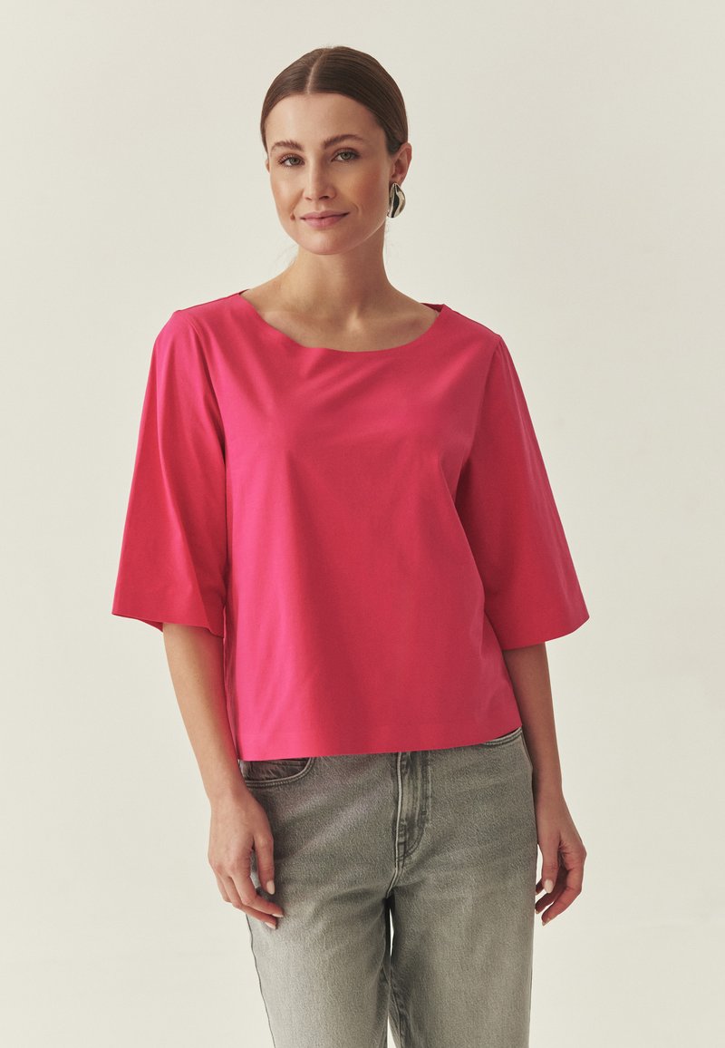 Haut rose à manches courtes avec un large col rond, fabriqué dans un tissu lisse. Associé à un jean taille haute gris clair, présentant une coupe décontractée.