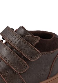 Sneaker in pelle marrone con due cinturini in Velcro, colletto imbottito, superficie texturizzata, cuciture a contrasto e suola in gomma marrone.