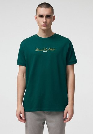 Młody mężczyzna ubrany w ciemnozieloną koszulkę z żółtym napisem "Diverse Sport Club New York", w jasnoszarych spodniach, stojący na tle jednolitej ściany.