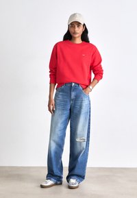 Red sweatshirt, light blue oversized jeans with a distressed detail, and metallic silver sneakers. Cream cap with logo accents.