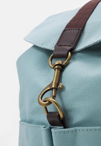 Helles blaue Stofftasche mit Messingverschluss, der an einem dunkelbraunen Riemen und einer Lederschlaufe befestigt ist, Nahaufnahme von der Tasche und dem Deckel.