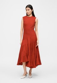 Γυναίκα που φορά ένα μίντι φόρεμα χωρίς μανίκια σε καμένο πορτοκαλί χρώμα με ασύμμετρο τελείωμα και λευκά μυτερά τακούνια, στέκεται σε λευκό φόντο.