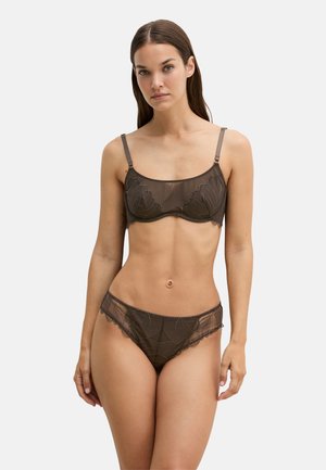 Vrouw draagt bijpassend bruin doorschijnend lingerie set met bloemenborduursel, staand tegen een effen lichte achtergrond.
