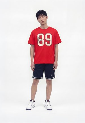 Een jonge man staat, gekleed in een rood T-shirt met nummer 89, marineblauwe korte broek met witte bies en witte sneakers tegen een effen witte achtergrond.