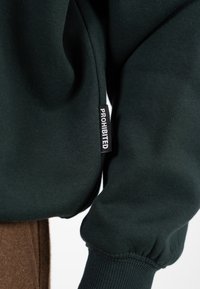 Primo piano di una manica di felpa verde scuro con etichetta nera che recita "PROIBITO" in lettere bianche.