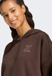 Donna sorridente con capelli intrecciati che indossa un cappuccio Adidas marrone scuro con il testo "three stripes studio" sul petto e piccoli orecchini a cerchio dorati.