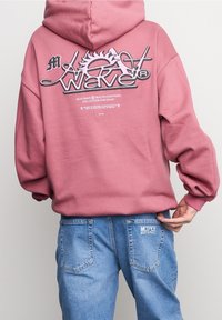 Sudadera rosa con un gran logotipo gráfico en la parte posterior, que presenta letras redondeadas y un motivo de sol. Combinada con jeans de mezclilla azul claro.