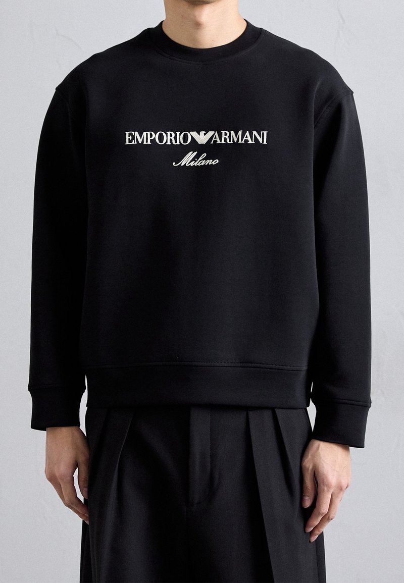 Musta collegepaita, joka on valmistettu puuvillasekoitteesta. Siinä on valkoisella brodeerattu "EMPORIO ARMANI Milano" -teksti, pyöreä kaula-aukko ja resorit hihansuissa.