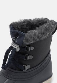 Acercamiento a una bota de invierno negra impermeable con un suave forro de felpa gris y un sistema de cordones, resaltando la textura y el ajuste seguro.