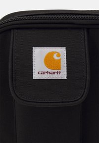 Bolsa de tela negra con un parche cuadrado blanco que muestra un logo amarillo y el texto "carhartt" en rojo. La bolsa tiene cierre de cremallera y superficie texturizada.