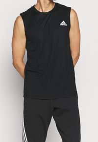 Haut de sport noir sans manches en tissu lisse, avec un logo Adidas blanc sur la poitrine gauche et de fines rayures aux épaules.