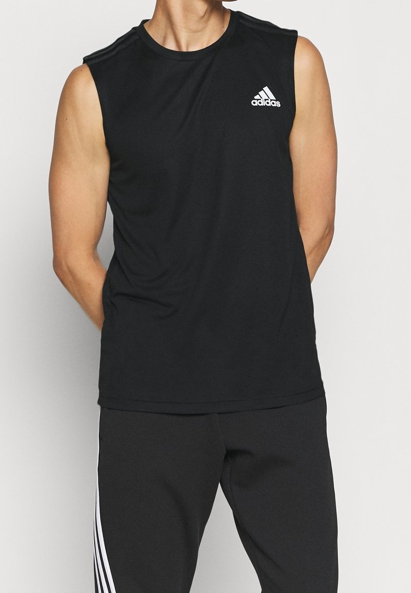 Haut de sport noir sans manches en tissu lisse, avec un logo Adidas blanc sur la poitrine gauche et de fines rayures aux épaules.