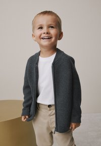 Cardigan gris tricoté avec une texture côtelée, fermeture éclair à l'avant, porté sur un t-shirt blanc. Associé à un pantalon beige, sur un fond neutre.