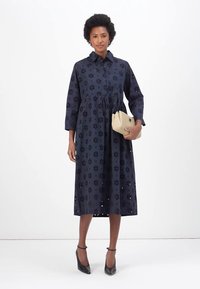 Abito lungo a maniche lunghe di color blu navy con motivi floreali ad occhiello, chiusura a bottoni davanti e vita arricciata. Il modello tiene una pochette beige e indossa tacchi neri.