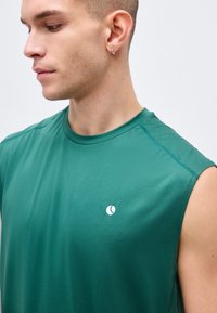 Νεαρός με κοντά μαλλιά και σκουλαρίκια στα αυτιά, φορόντας πράσινο αμάνικο αθλητικό πουκάμισο με ένα μικρό λευκό λογότυπο στο στήθος.