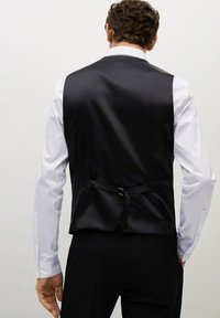 Gilet di satin nero con una texture liscia, caratterizzato da una cinghia posteriore singola e un colletto a punta, indossato sopra una camicia bianca a maniche lunghe.