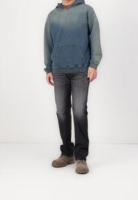 Hombre vestido con una sudadera azul descolorida, jeans gris oscuro y zapatos beige con cordones, de pie contra un fondo blanco liso.