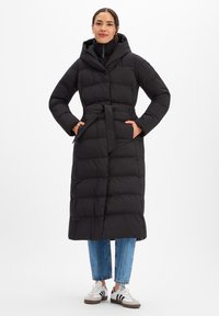 Lange zwarte donsjas met capuchon, taille met riem, ritskraag en zijzakken, gecombineerd met een blauwe spijkerbroek en witte sneakers.