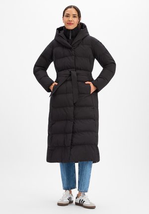 Lange zwarte donsjas met capuchon, taille met riem, ritskraag en zijzakken, gecombineerd met een blauwe spijkerbroek en witte sneakers.