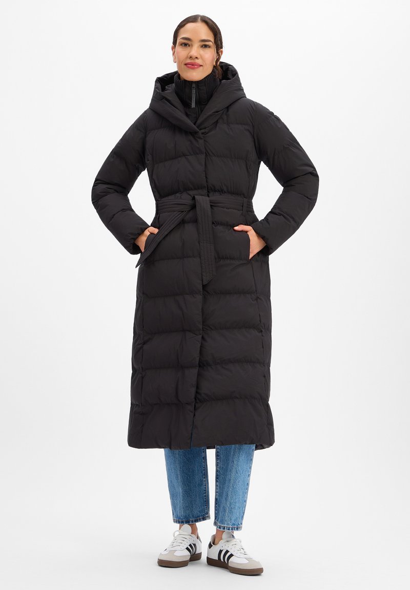 Lange zwarte donsjas met capuchon, taille met riem, ritskraag en zijzakken, gecombineerd met een blauwe spijkerbroek en witte sneakers.