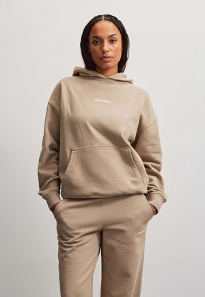 Kvinde iført matchende beige hættetrøje og joggingbukser med hænderne i lommerne, stående mod en ensfarvet lys baggrund.