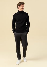 Schwarzer Rollkragenpullover, graue maßgeschneiderte Hose und weiße Sneakers. Lockige Haare, entspannte Pose, stehend mit den Händen in den Taschen vor einem hellen Hintergrund.
