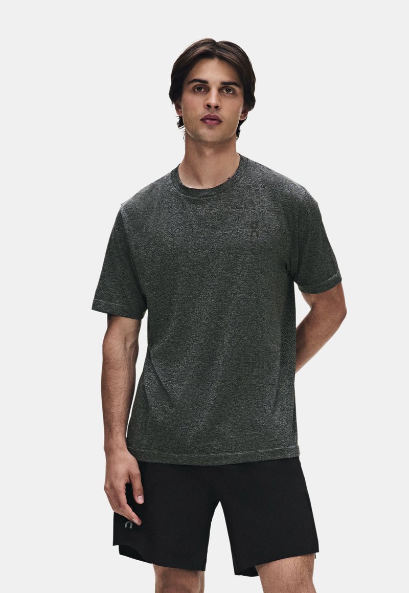 Jeune homme debout, face à l'appareil, portant un T-shirt gris foncé à manches courtes et un short noir sur un fond clair uni.