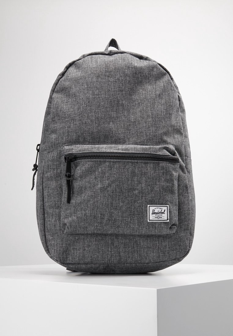 Sac à dos en tissu gris avec poche avant zippée et fermeture éclair noire, placé sur une surface blanche sur fond gris clair uni.