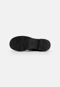 Black rubber shoe sole featuring a textured pattern with grooves and ridges for traction. Smooth edges and a circular logo visible.
