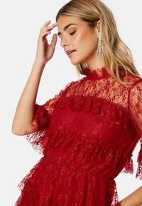 Abito rosso in pizzo con collo alto, maniche arricciate e dettagli a strati, decorato con un motivo in pizzo floreale su tutta la superficie.