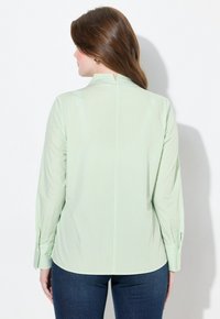 Camicia a righe verde chiaro con colletto alla coreana e maniche lunghe. Presenta un piccolo spacco nella parte posteriore del collo e maniche con polsini.