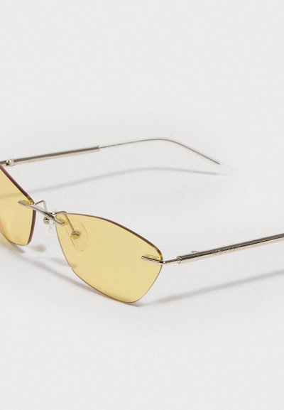 Keltaisenväriset aurinkolasit, joissa on ohut hopeinen metallirunko ja kulmikkaat linssit. Varustettu nenävarrella ja hienovaraisesti kaarevilla temppeleillä.