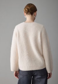 Lys rosa, fluffy genser med lange ermer og rund hals, brukt med mørkegrå bukser. Myk tekstur, minimalistisk design.