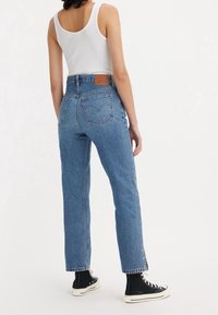 Mujer con jeans de tiro alto azules, camiseta sin mangas blanca y zapatillas negras de caña alta, de pie de espaldas frente a la cámara sobre un fondo blanco.