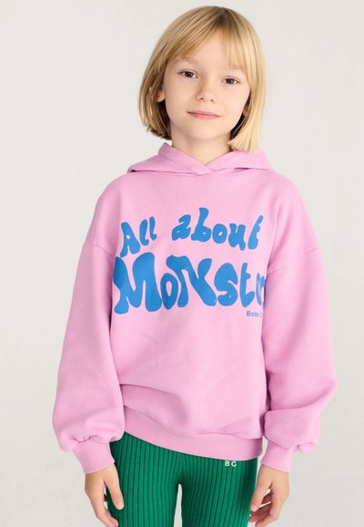 Enfant blond portant un sweat à capuche rose avec un texte bleu "All about Monsters" et un pantalon côtelé vert, debout sur un fond neutre.
