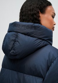 Osoba s tmavými kudrnatými vlasy, oblečená v námořnickomodré péřové bundě s kapucí, která má na kapuci jemně vyšitý text.