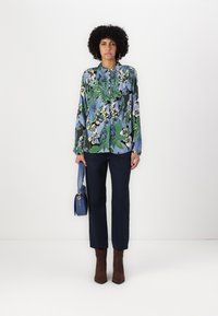 Floral blouse with green, blue, and black patterns; long sleeves; collared. Paired with navy trousers and brown heeled boots. Holding a blue handbag.