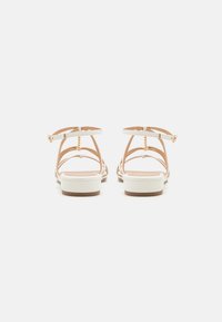 Lys beige stroppsandaler med hvite accenter og flate såler. Har gulltone spenne og en detaljert bakdesign.
