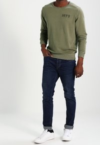 Olivgrön sweatshirt med rund halsringning och "1973" tryckt på bröstet; matchad med mörkblå jeans och vita sneakers.