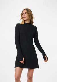 Žena v čiernych pletených šatách s dlhým rukávom a otvorom na palce, stojaca pred bielym pozadím, mierne otočená bokom.