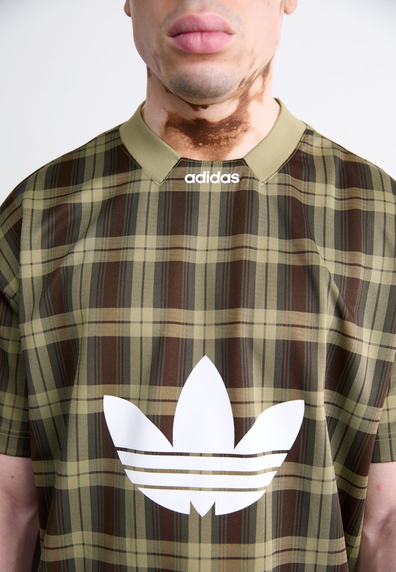 Brązowa i zielona koszulka w kratę z białym logo Adidasa i trzema paskami z przodu. Posiada zielany kołnierzyk i krótki rękaw.