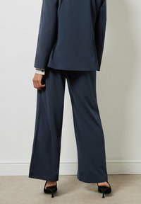 Navy blauwe op maat gemaakte kostuum met wijde pijpen. Heeft een gestructureerde afwerking en een subtiele glans. Zwarte hoge hakken complementeren de outfit.