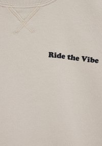 Svetlo béžová látka s čiernym nápisom "Ride the Vibe", zobrazuje detail stehov výstrihu na vrchu.