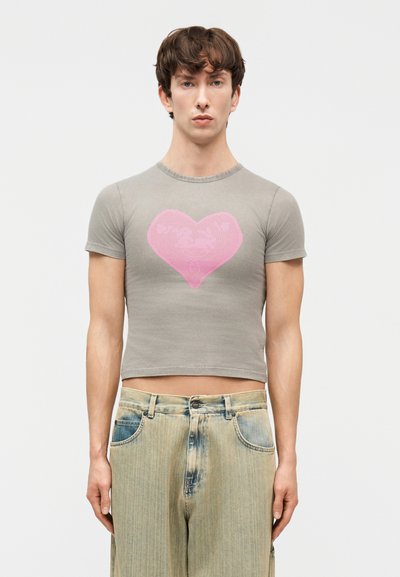 Νεαρός άνδρας που φοράει εφαρμοστό γκρι κοντομάνικο μπλουζάκι με μεγάλο ροζ καρδιά σχεδιασμό, συνδυασμένο με ξεθωριασμένο wide-leg τζιν.