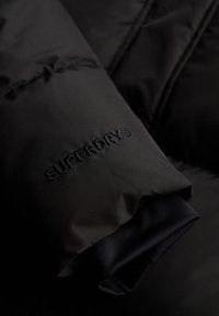 Zwarte gewatteerde mouw van een jack met het geborduurde logo 'SUPERDRY'. Glad textuur met een matte afwerking en gelaagde stofdetails.